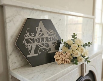 Può includere: Una piastrella esagonale grigia con il nome "Anderson" e la lettera "A" in un carattere decorativo. Un cartello di legno con la scritta "Mr. & Mrs." è accanto a un bouquet di rose bianche e verde. Su un camino di marmo.