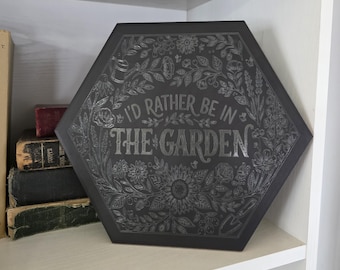 Può includere: Un cartello esagonale nero con la frase "I'd rather be in the garden" in caratteri bianchi. Il cartello è circondato da un disegno floreale. È esposto su una mensola bianca con libri.