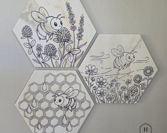 Può includere: Tre piastrelle esagonali con disegni a tratto nero di api e fiori. Una piastrella mostra un'ape con trifoglio e lavanda, un'altra con un'ape che vola sopra i fiori e l'ultima con un'ape e un favo. Le piastrelle sono bianche con un motivo marmorizzato.