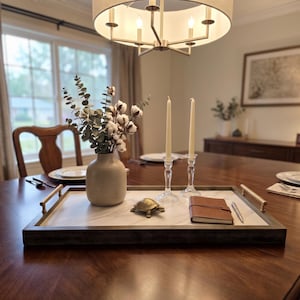 May include: A rectangular serving tray with a white marble-patterned base, dark wood frame, and gold-toned metal handles. On the tray sits a beige ceramic vase with cotton stems, two clear glass candlesticks with taper candles, a small brass turtle, and a brown leather-bound notebook with a silver pen.
