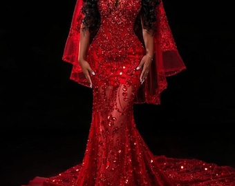 May include: A red, floor-length gown with a plunging neckline and a sheer, embellished skirt. The dress is covered in sequins and features a long train. A matching red veil drapes over the shoulders. The background is black.