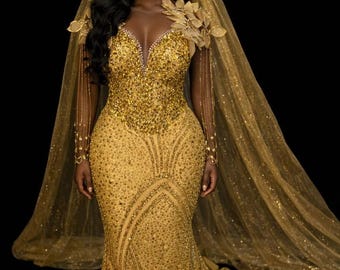 May include: A golden, beaded gown with a plunging neckline and a flowing train. The dress features intricate beadwork and is paired with a matching veil. The sleeves are long and adorned with delicate bead strands. The overall style is elegant and formal.