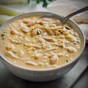 Puede incluir: Un cuenco de chili de pollo blanco cremoso con frijoles, maíz y hierbas verdes. Una cuchara plateada descansa en el cuenco, lista para servir. El cuenco azul claro está sobre una superficie gris.