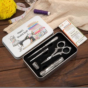 May include: A white metal sewing kit tin with the text Marie's Sewing Stuff and a sewing machine illustration. Inside, a black foam insert holds silver-toned metal sewing tools including scissors, a thimble, a needle case, and two awls.