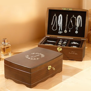 May include: A wooden jewelry box with a hinged lid, open to reveal necklaces, rings, and bracelets. The box is brown with a gold clasp and a floral design on the lid. The open box displays various necklaces and rings.