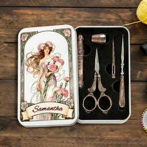 May include: A rectangular metal storage tin featuring an illustration of a woman with flowers and the name Samantha on a banner. The open tin contains a set of bronze-colored sewing tools including scissors, a thimble, a needle case, and an awl.