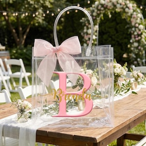 May include: Clear tote bag with a pink bow and a large pink letter "B" with the name "Brenda" in gold script. The bag has a clear handle and is sitting on a wooden table.