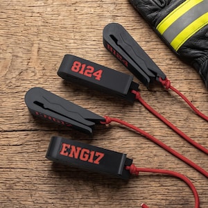 May include: Four black plastic glove clips with red cord attachments. Two clips feature the red text 8124 and two clips feature the red text ENG17. The clips have a jagged interior grip design and are positioned near a black leather glove.