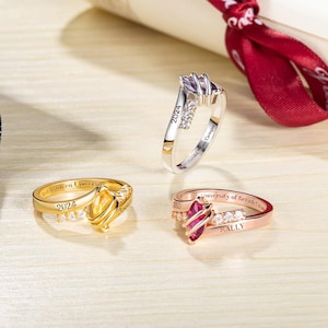 May include: Three graduation rings with gemstones. One is gold with a yellow stone, one is silver with a purple stone, and one is rose gold with a red stone. Each ring has the year 2024 or 2025 engraved on it, along with a name and university.