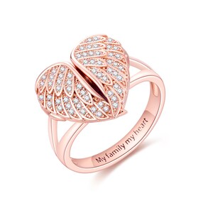 May include: Rose gold ring featuring a heart-shaped design with two open wings, encrusted with small, clear gemstones. The ring band is engraved with the words "My family my heart".