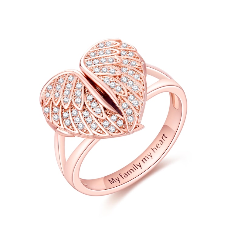 May include: Rose gold ring featuring a heart-shaped design with two open wings, encrusted with small, clear gemstones. The ring band is engraved with the words "My family my heart".
