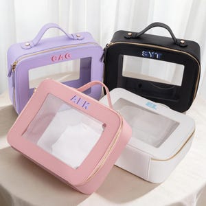 May include: Four cosmetic bags in pastel colors: lavender, pink, black, and white. Each bag has a clear window, a top handle, and a gold zipper. The bags have initials printed on them: GAG, AK, SYT, and EL.