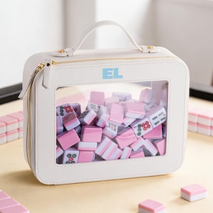 May include: A white, square Mahjong tile carrying case with a clear window displaying pink and white tiles. The case has a top handle, gold zipper, and the initials "EL" in the upper right corner. Additional tiles are scattered around the case.