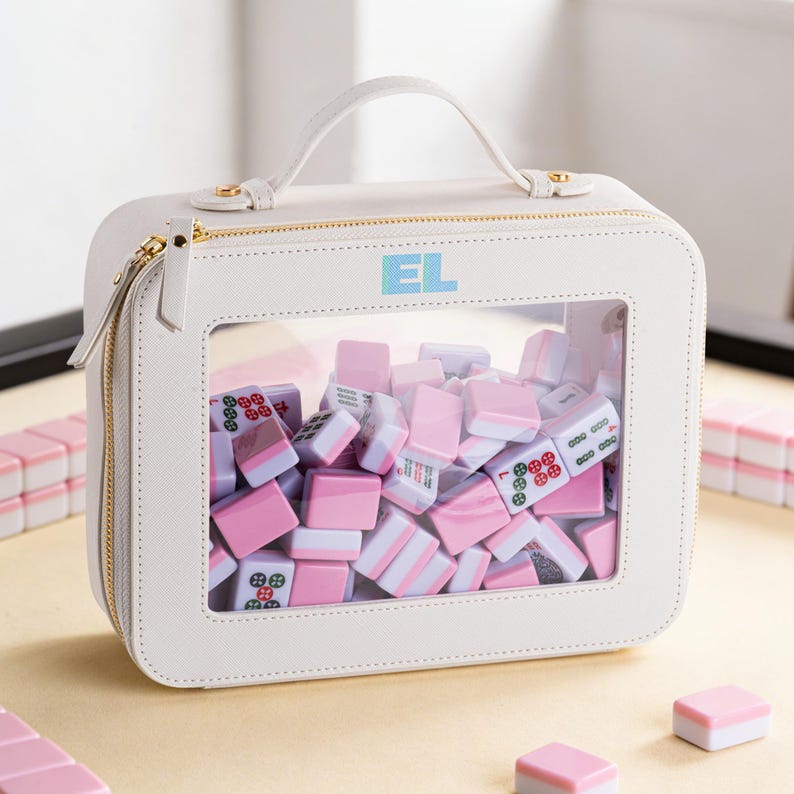 May include: A white, square Mahjong tile carrying case with a clear window displaying pink and white tiles. The case has a top handle, gold zipper, and the initials "EL" in the upper right corner. Additional tiles are scattered around the case.