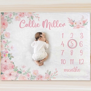 Op de afbeelding: Een baby ligt op een witte deken met roze bloemaccenten en de naam "Callie Miller" in roze letters. De deken heeft de cijfers 1-12, met het cijfer 5 omcirkeld, wat de leeftijd van de baby in maanden aangeeft.