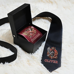 Puede incluir: Una caja de regalo negra con un cinturón de cuero rojo en su interior, con un emblema dorado y el nombre "Samuel". Una corbata negra con un emblema a juego y el nombre "Oliver" está junto a la caja. Sobre una superficie blanca y esponjosa.