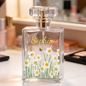 May include: A rectangular clear glass perfume bottle with a silver-toned spray nozzle and a transparent cap. The front features the name Sophia in yellow script, surrounded by a printed design of white daffodils with yellow centers and small butterflies.