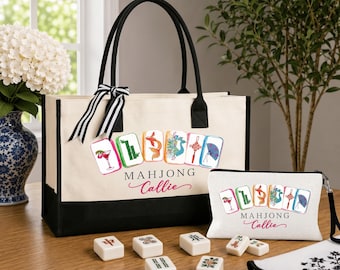 Puede incluir: Un bolso de lona color crema y un estuche con cremallera a juego, ambos con ribete negro y un gráfico de seis fichas de mahjong con el texto "MAHJONG" y el nombre "Callie" en letra rosa. Varias fichas de mahjong están esparcidas cerca.