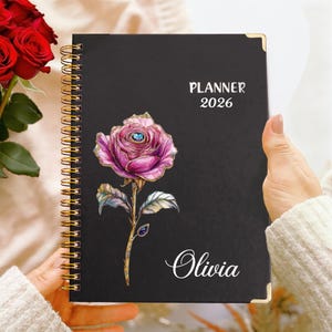 Pode incluir: Agenda preta para 2026 com encadernação em espiral e cantos dourados. A capa apresenta uma ilustração de rosa rosa com detalhes dourados, o nome "Olivia" em escrita cursiva e o texto "PLANNER 2026" em branco.
