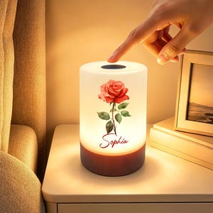May include: A cylindrical table lamp with a white frosted shade featuring a red rose illustration and the name Sophia printed in red script. The lamp has a dark wood-grain base and a circular touch-sensitive power button on the top surface.