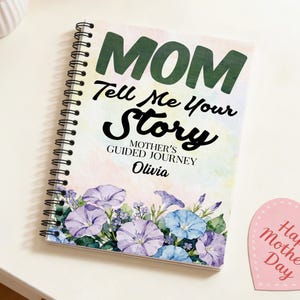 以下が含まれることがあります： 緑と黒で「MOM Tell Me Your Story」と書かれたスパイラル綴じノート。表紙には紫と青の色合いの水彩画の花が描かれています。このノートは、オリビア向けにパーソナライズされた「Mother's Guided Journey」です。