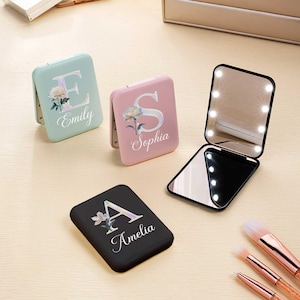 May include: Compact, personalized makeup mirrors in mint green, pink, and black. Each mirror features a floral design with a letter and a name: "Emily", "Sophia", and "Amelia". One mirror is open, revealing LED lights.