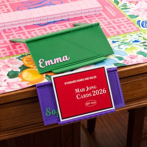 May include: A green plastic Mah Jong rack with the name Emma in pink lettering, and a purple plastic Mah Jong rack holding a red card with white text that reads Standard Hands and Rules Mah Jong Cards 2026 89th Year, positioned on a table.