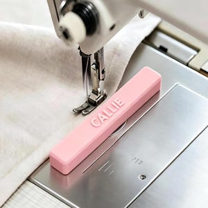Op de afbeelding: Een rechthoekige, lichtroze magnetische naaigids voor naaimachines met de naam CALLIE in reliëf in hoofdletters op het bovenoppervlak. De gids is geplaatst op de metalen steekplaat van een naaimachine naast een stuk lichte stof.