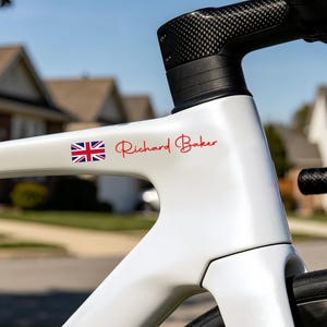 May include: Close-up of a white bicycle frame with a black carbon fiber handlebar. The frame features a Union Jack flag sticker and the name "Richard Baker" in red script. The background is blurred, showing a residential area.