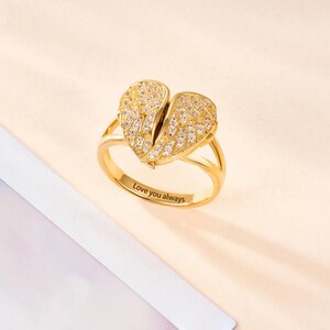 May include: A gold-colored ring with a heart-shaped design, split in the middle, resembling angel wings. The wings are encrusted with small, clear stones. The ring band has a split design and the words "Love you always" are engraved on the band.