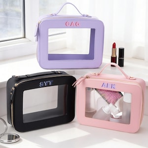 May include: Three cosmetic bags in lavender, black, and pink. Each bag has a clear window, a top handle, and a zipper closure. The bags are personalized with initials. The pink bag contains beauty products.