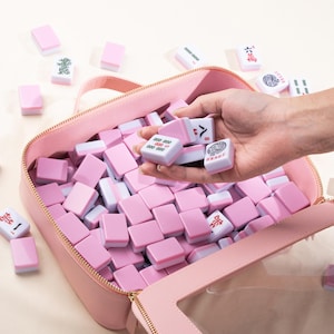 May include: A pink zippered bag filled with pink and white Mahjong tiles. Some tiles are held in a hand, while others are scattered around the bag. The tiles have various symbols and characters.