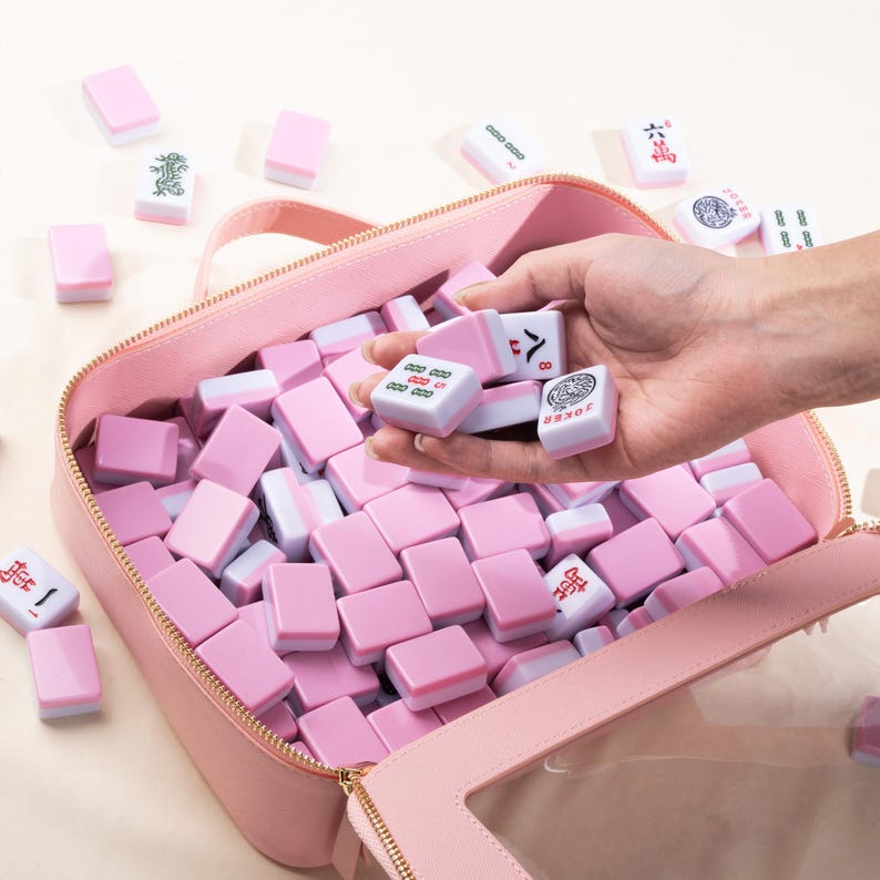May include: A pink zippered bag filled with pink and white Mahjong tiles. Some tiles are held in a hand, while others are scattered around the bag. The tiles have various symbols and characters.