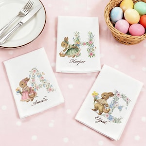 May include: White Easter-themed decorative towels with bunny illustrations and floral letter designs. Each towel features a different name: Harper, Amelia, and Elijah. A basket of colorful Easter eggs is in the background.