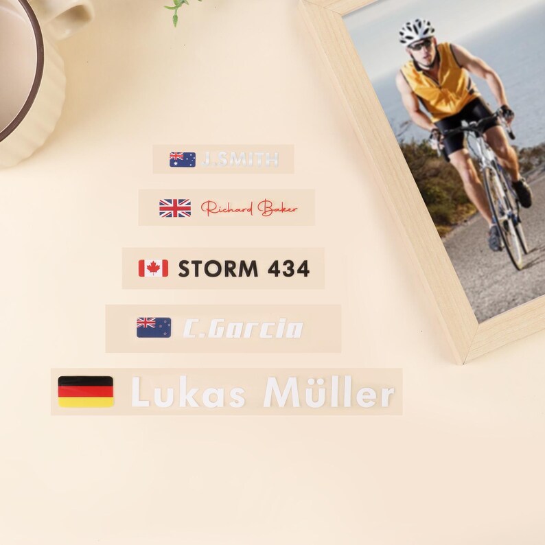 May include: A collection of name tags with country flags and names. The tags include the flags of Australia, the United Kingdom, Canada, and Germany. The names are J. Smith, Richard Baker, STORM 434, C. Garcia, and Lukas M&uuml;ller. A framed picture of a cyclist is in the background.