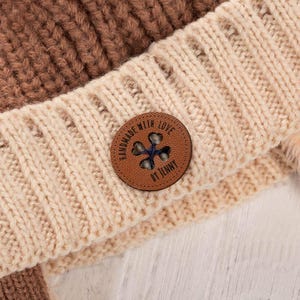May include: Close-up of a knitted beanie in shades of brown and cream. A round, brown leather patch with the text "HANDMADE WITH LOVE BY JENNY" and a cross-shaped design is sewn onto the beanie. The beanie is made of a thick, textured yarn.