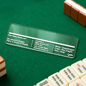Puede incluir: Una hoja de trucos de Mahjong de acrílico transparente con tinte verde, que muestra instrucciones y reglas del juego. El texto incluye "Play Right, Walls Left" y "Charleston". La hoja está sobre una superficie de fieltro verde, con fichas de Mahjong en el fondo.