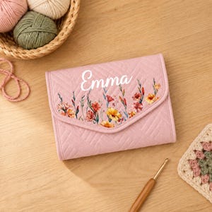 May include: A light pink quilted fabric case featuring a chevron pattern and a floral print with the name Emma written in white script. The rectangular case has a flap closure and is positioned near yarn balls and a wooden crochet hook.