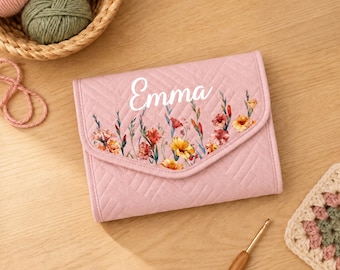 May include: A light pink quilted fabric case featuring a chevron pattern and a floral print with the name Emma written in white script. The rectangular case has a flap closure and is positioned near yarn balls and a wooden crochet hook.
