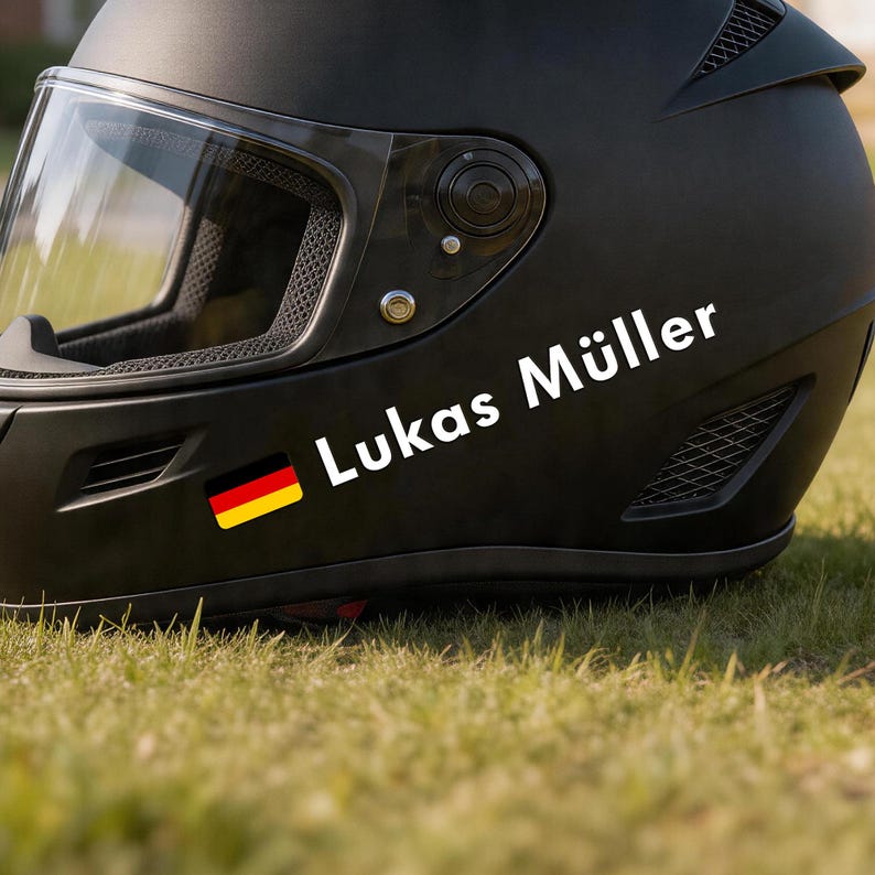 May include: A black motorcycle helmet with a clear visor. The name "Lukas M&uuml;ller" is printed in white, along with a small German flag sticker. The helmet is resting on green grass.