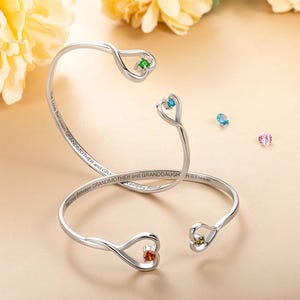 May include: Two silver-toned bracelets with heart-shaped ends, each holding a gemstone. One bracelet has the inscription "The Love Between GRANDMOTHER and GRANDDAUGHTER is Forever". Gemstones are green, blue, orange, and yellow.