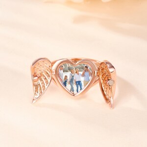 May include: Rose gold heart-shaped locket ring with wing details. The locket opens to reveal a family photo. The ring is displayed on a light peach background. The wings are open on either side of the heart.
