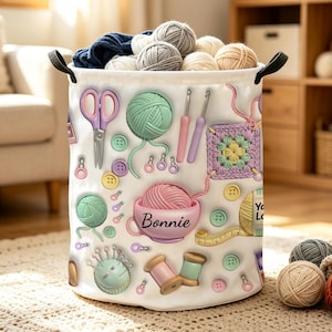 May include: A white cylindrical fabric storage basket filled with various yarn balls. The exterior features a printed pattern of craft supplies including scissors, crochet hooks, buttons, a pincushion, and a pink bowl labeled Bonnie.