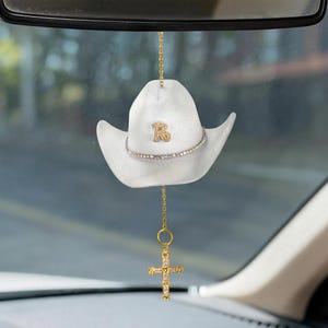 May include: White cowboy hat car charm with a gold-colored letter "R" and a rhinestone band. A gold chain suspends a gold cross charm with clear rhinestones. The charm hangs from a car's rearview mirror.