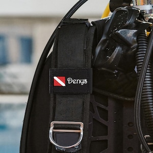 Peut inclure: Un gilet stabilisateur de plongée noir doté d'une sangle d'épaule rembourrée avec un écusson rectangulaire. L'écusson affiche une icône de drapeau de plongée rouge et blanc à côté du nom Denys brodé en blanc et un anneau en D métallique.