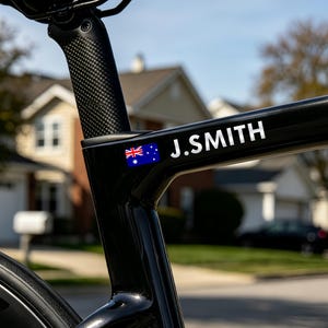 May include: Close-up of a black bicycle frame with the name "J.SMITH" in white lettering. An Australian flag sticker is visible. The bike is outdoors with houses and trees in the background.