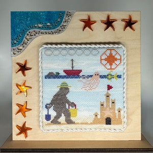 May include: A framed cross-stitch design featuring a cartoon monster holding a bucket on a beach with a sandcastle, a boat, and a ghost. The frame is made of wood and has a blue and white patterned border. There are five orange stars on the frame.