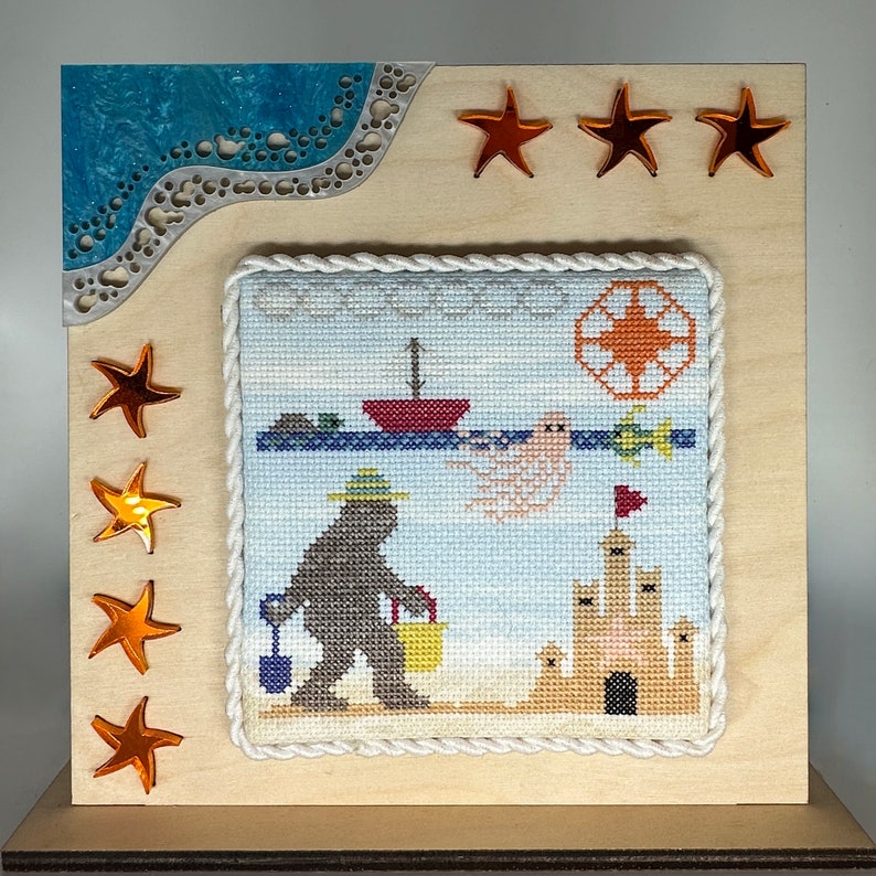 May include: A framed cross-stitch design featuring a cartoon monster holding a bucket on a beach with a sandcastle, a boat, and a ghost. The frame is made of wood and has a blue and white patterned border. There are five orange stars on the frame.