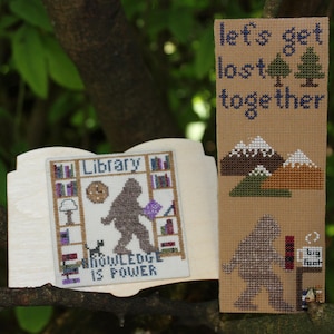 Può includere: Due pezzi a punto croce. Uno raffigura un Bigfoot in una biblioteca con la scritta "KNOWLEDGE IS POWER". L'altro dice "let's get lost together" con montagne e alberi. Entrambi su sfondo in legno chiaro.