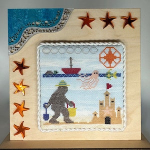 May include: A cross-stitch embroidery framed in a light wood frame with a white rope border. The embroidery depicts a brown bear wearing a blue hat and carrying a bucket, a sandcastle, a seagull, and a red boat. The frame is decorated with orange star-shaped embellishments.